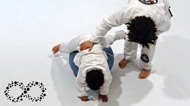 Imagem mostra uma aula de JiuJitsu capturando uma interação entre a instrutora e uma criança pequena, ambas vestidas com o tradicional Gi (kimono) da marca portuguesa ACADEMIA DE JIUJITSU OCUPACIONAL - POR PAULA FIGUEIREDO®.