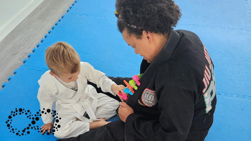 Imagem mostra uma aula de JiuJitsu capturando uma interação entre a instrutora e uma criança pequena, ambas vestidas com o tradicional Gi (kimono) da marca portuguesa ACADEMIA DE JIUJITSU OCUPACIONAL - POR PAULA FIGUEIREDO®.