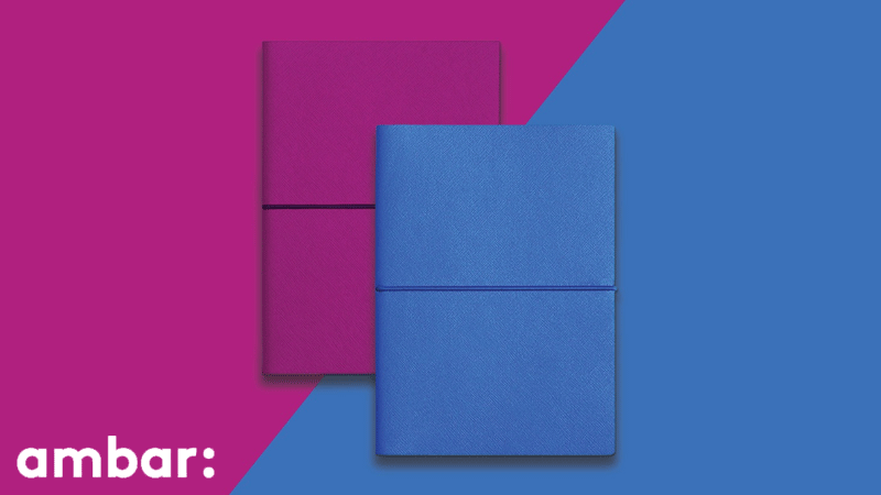 Dois cadernos, um azul e outro cor-de-rosa, sobre um fundo dividido entre azul e cor-de-rosa, com o logótipo da marca portuguesa AMBAR®.