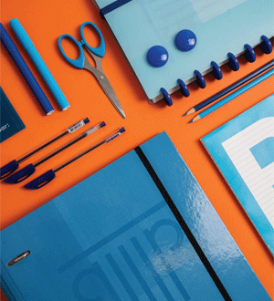 Material escolar e de escritório em tons de azul, incluindo cadernos, lápis, canetas e tesoura, disposto sobre uma superfície cor de laranja da marca portuguesa AMBAR®.