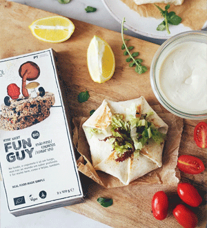 Embalagem de hambúrguer vegetal biológico Fun Guy (de feijão preto e cogumelos) da marca portuguesa Beatroot, acompanhada por um wrap preparado, rodelas e pedaços de limão, tomates cherry e um frasco com molho/natas ácidas de caju.