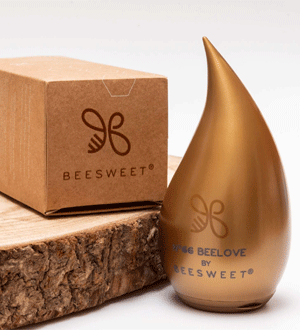Embalagem dourada de mel da marca portuguesa BEESWEET®, em forma de gota, junto a uma caixa de cartão sobre uma rodela de madeira.