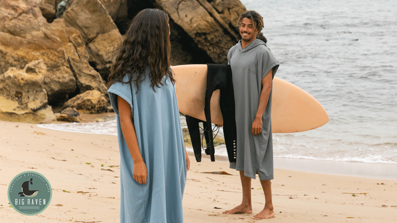 Duas pessoas na praia a usar ponchos da marca portuguesa BIG RAVEN PORTUGAL, junto a uma prancha de surf e a uma zona rochosa junto ao mar.
