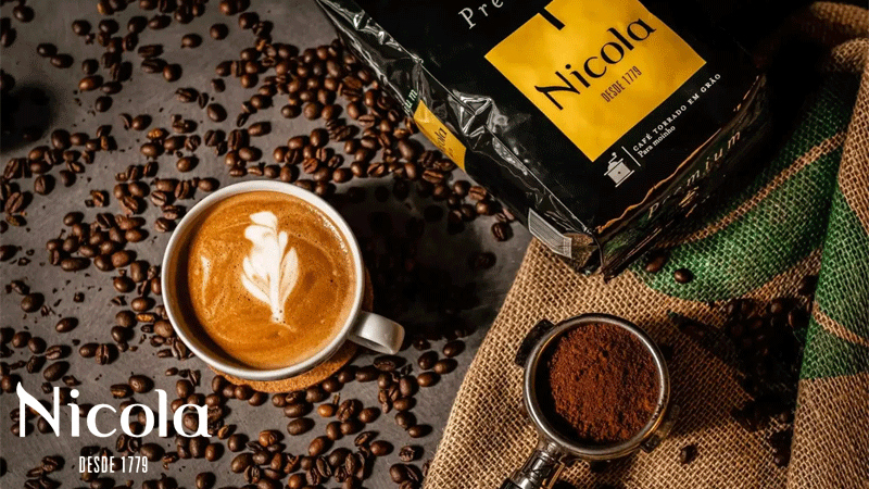 Café da marca portuguesa CAFÉ NICOLA® servido numa chávena, rodeado por grãos de café, café moído e uma embalagem da marca, com a assinatura “Desde 1779”.