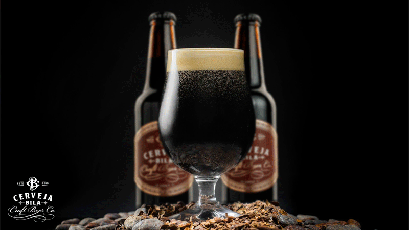 Copo de cerveja escura da marca portuguesa CERVEJA BILA® com espuma cremosa, em primeiro plano, com duas garrafas desfocadas ao fundo sobre cevada e malte.