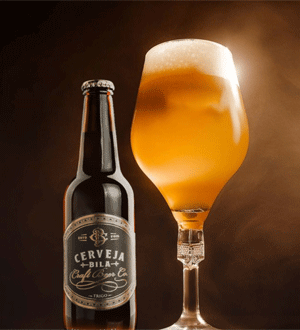 Garrafa da marca portuguesa CERVEJA BILA® ao lado de um copo de cerveja dourada com espuma cremosa, sobre fundo escuro e iluminação quente.