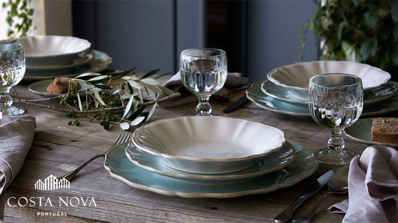 Mesa rústica posta com pratos e travessas da marca portuguesa COSTA NOVA® em tons suaves de verde e branco, acompanhados por copos, talheres e ramos de oliveira.