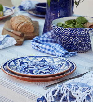 Mesa posta com loiça da marca portuguesa COSTA NOVA® em tons de azul e branco, com pratos decorados, taça, jarro, pão e têxteis de padrão xadrez.