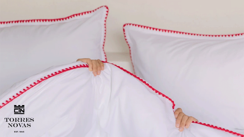 Roupa de cama branca da marca portuguesa CTN TORRES NOVAS EST. 1845®, com almofadas e edredão rematados por detalhe decorativo vermelho.