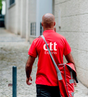 Courier from the Portuguese brand CTT®, seen from behind, wearing a red polo shirt and carrying a red shoulder bag along a cobbled street.