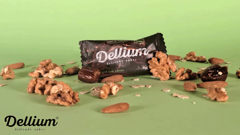 Barrita da marca portuguesa DELLIUM, DELICADO SABOR® com sabor a maçã e cacau, apresentada entre nozes, amêndoas, tâmaras e flocos de aveia sobre fundo verde.