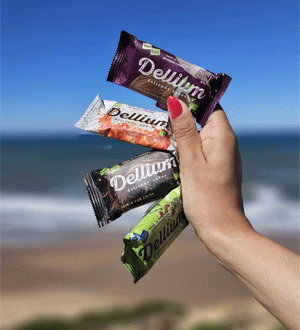 Mão a segurar quatro barritas da marca portuguesa DELLIUM, DELICADO SABOR® junto ao mar, com diferentes embalagens e sabores.