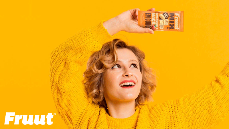 Mulher sorridente de camisola amarela segura uma embalagem laranja da marca portuguesa FRUUT® acima da cabeça, sobre fundo amarelo.