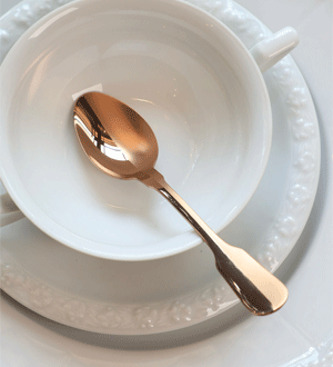 Colher de chá de metal com acabamento em tom de cobre da marca portuguesa Herdmar, colocada dentro de uma xícara branca clássica.