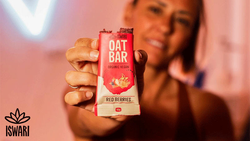 Mulher segura uma barra de aveia com frutos vermelhos da marca portuguesa ISWARI®, em primeiro plano, num ambiente de tons quentes.