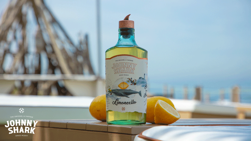Garrafa de limoncello artesanal português da marca portuguesa Johnny Shark, rodeada de limões frescos cortados.