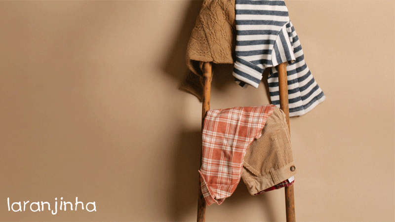 Conjuntos de roupa infantil da marca portuguesa Laranjinha pendurados numa pequena escada de madeira.