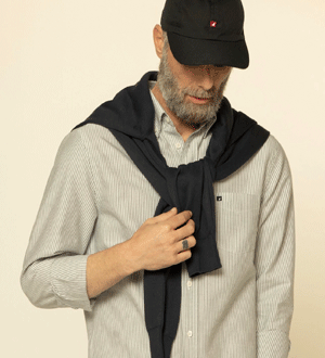 Peças de vestuário de estilo clássico e casual da marca portuguesa Mike Davis. Peças versáteis ideais para o dia a dia. O conjunto inclui uma camisa de botões com riscas finas, um suéter atado sobre os ombros e um boné de preto.