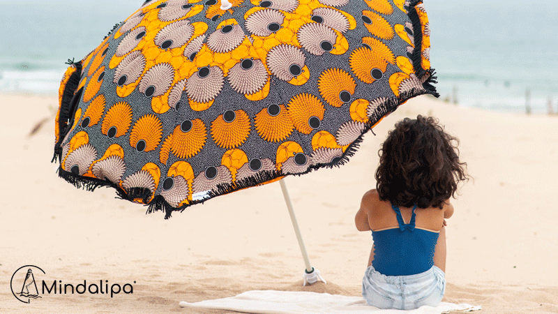 Guarda-sol da marca portuguesa MINDALIPA® com padrão africano em tons de laranja, preto e bege, numa praia, junto a uma mulher sentada numa toalha.