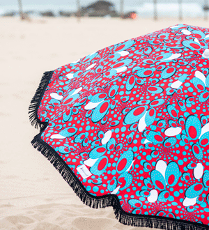 Pormenor de guarda-sol da marca portuguesa MINDALIPA® com padrão colorido em vermelho, azul e branco e acabamento com franjas pretas, numa praia.