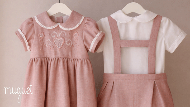 Roupas infantis clássicas da marca portuguesa Muguet, nomeadamente, um vestido rosa com bordados e um conjunto de jardineira, ambos em tons rosa velho e branco.