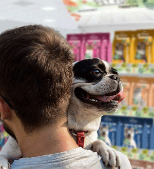 Pessoa com um cão ao colo numa loja de produtos para animais, com embalagens de alimentação animal ao fundo, da marca portuguesa PETMAXI®.