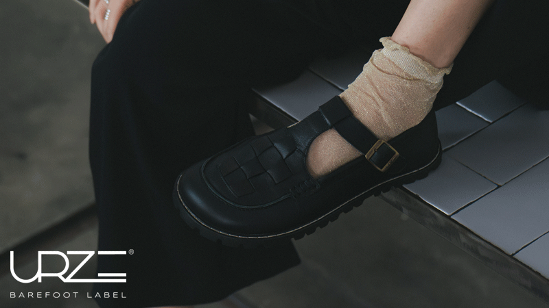 Sapato barefoot preto da marca portuguesa URZE BAREFOOT LABEL® com fivela lateral e detalhe entrançado, usado com meia em tom bege.