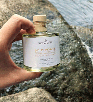 Mão a segurar um frasco de Body Power da marca portuguesa VEGBEAUTY® junto à água e a rochas, num ambiente natural.