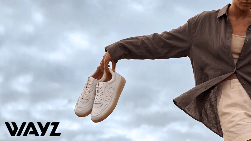 Pessoa de camisa castanha a segurar sapatilhas brancas com solas bege da marca portuguesa WAYZ®, com um céu nublado em fundo e o logótipo no canto inferior esquerdo.