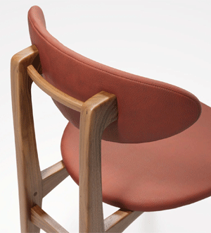 Cadeira em madeira maciça da marca portuguesa WEWOOD PORTUGUESE JOINERY®, com assento e encosto estofados em pele de tom terracota, vista em detalhe sobre fundo claro.