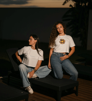 Duas mulheres vestem T-shirts brancas da marca portuguesa WHITEVULK® com calças de ganga, sentadas num espaço exterior de ambiente contemporâneo.