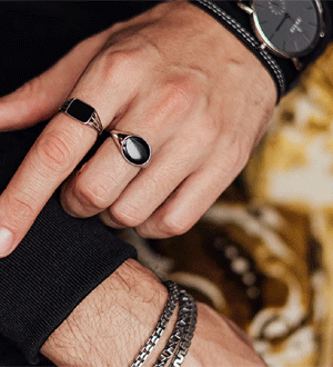 Seleção de acessórios masculinos elegantes, destacando anéis de sinete com pedras pretas, um relógio minimalista e uma pulseira de elos da marca portuguesa Zarco.